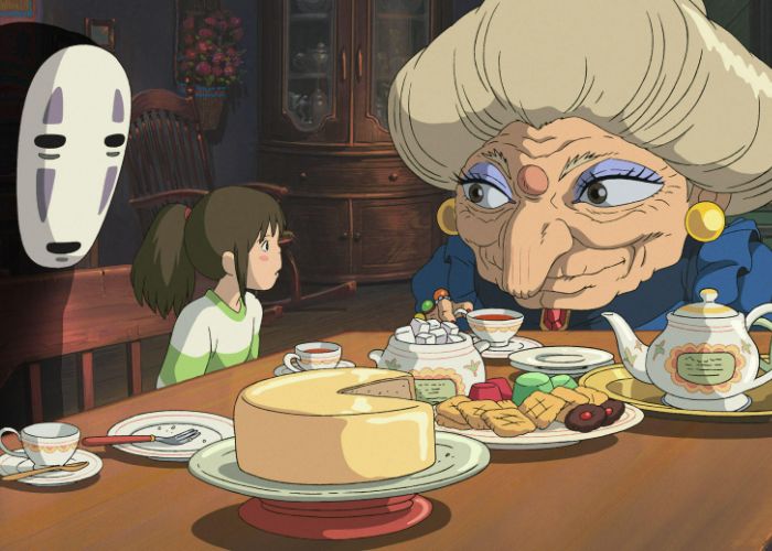 Chihiro and No-Face sitting at a table with Zeniba. In front of them, the table is covered in cakes, biscuits, and tea.
